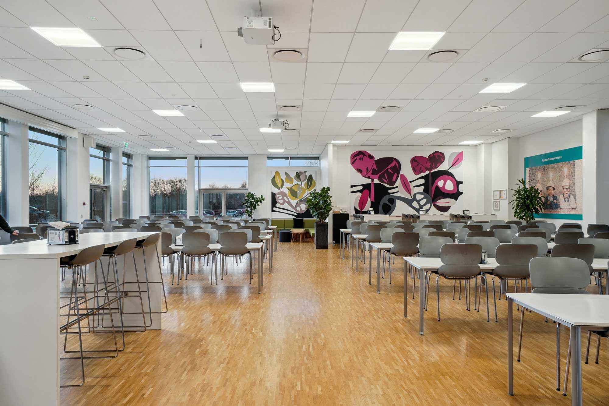 En stor fællesområde med højt loft og moderne møbler, inkluderende borde, stole og plantager. Der er et stort kunstværk på væggen og store vinduer med udsigt til byen.