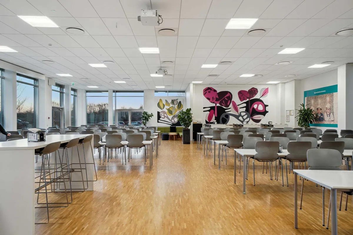 En stor fællesområde med højt loft og moderne møbler, inkluderende borde, stole og plantager. Der er et stort kunstværk på væggen og store vinduer med udsigt til byen.