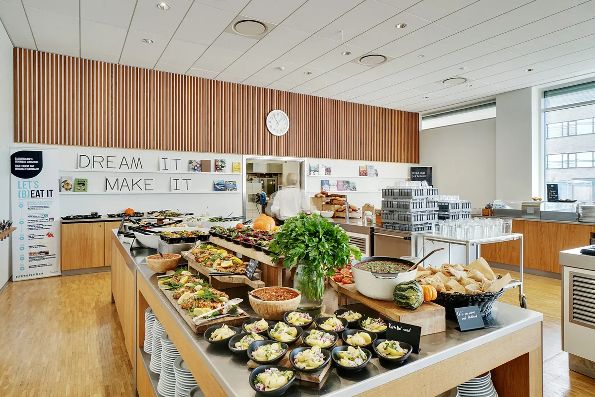 Buffet med forskellige retter og salater i en moderne og fleksibel kontorrestaurant med lyse loftspaneler og træbeklædning på væggen