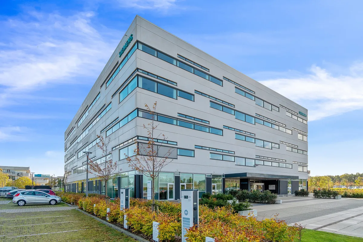 Siemens bygning med flere etager og moderne design. Foran bygningen er der parkeringsplads og grønne planter. Der er også biler parkeret på siden af bygningen