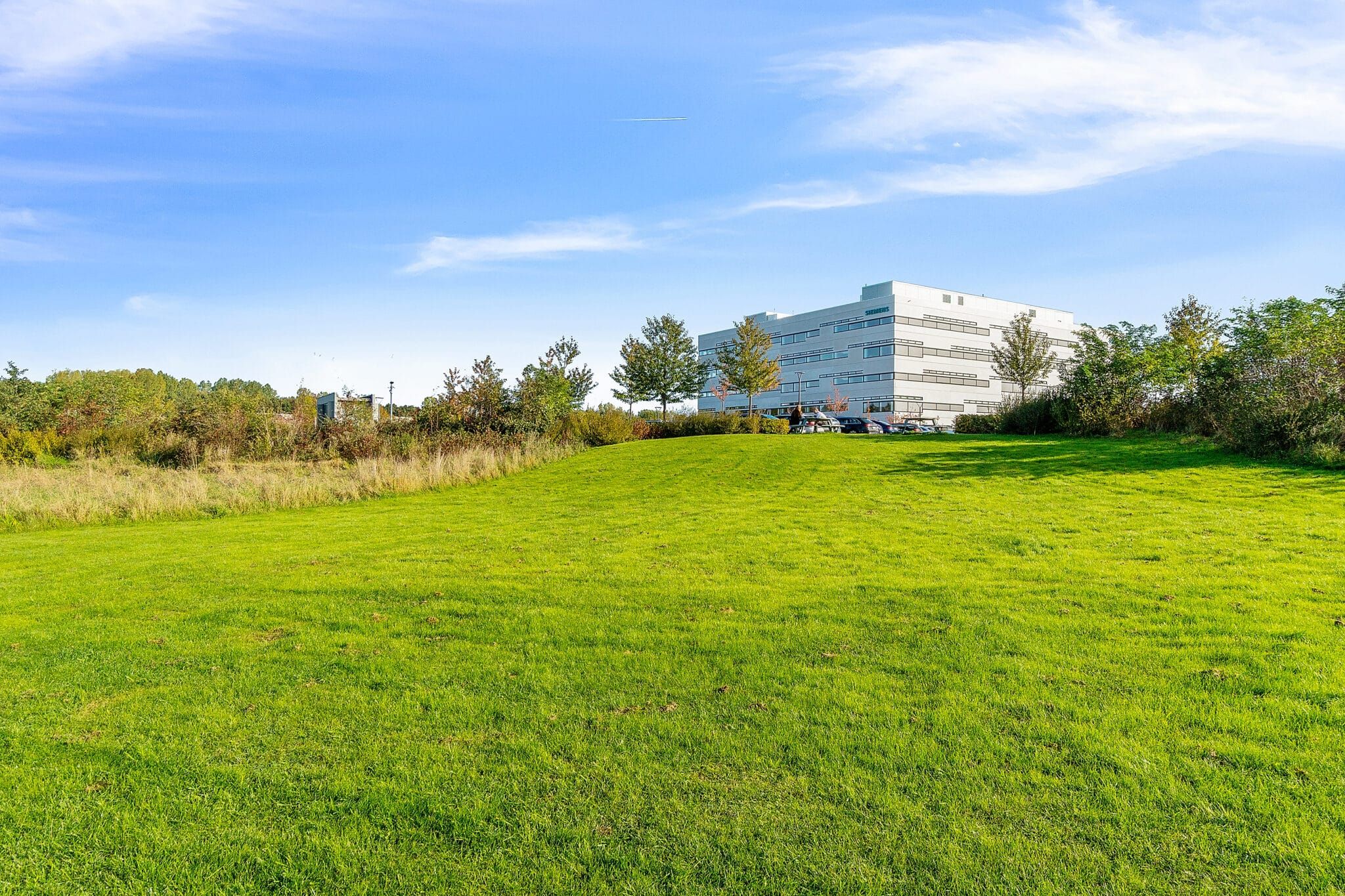 Et stort hvidt kontorhuse med flere vinduer og parkeringsplads foran bygningen. Omkring huset er der en stor grøn græsplæne med træer og buske.