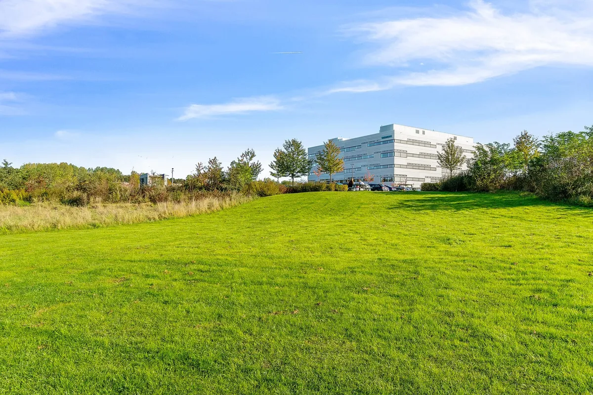 Et stort hvidt kontorhuse med flere vinduer og parkeringsplads foran bygningen. Omkring huset er der en stor grøn græsplæne med træer og buske.