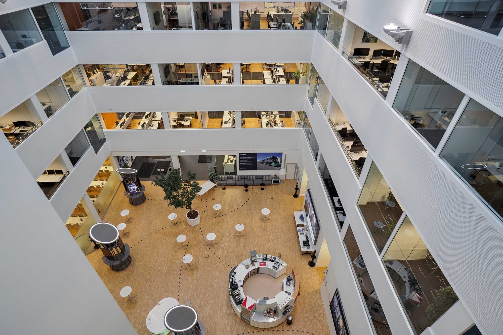 Kontorbygning med åben atrium, lyse gulve, runde borde, mange vinduer og møbler samt en stor plante i midten.