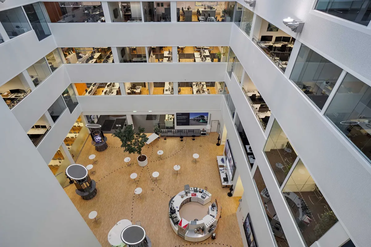 Kontorbygning med åben atrium, lyse gulve, runde borde, mange vinduer og møbler samt en stor plante i midten.