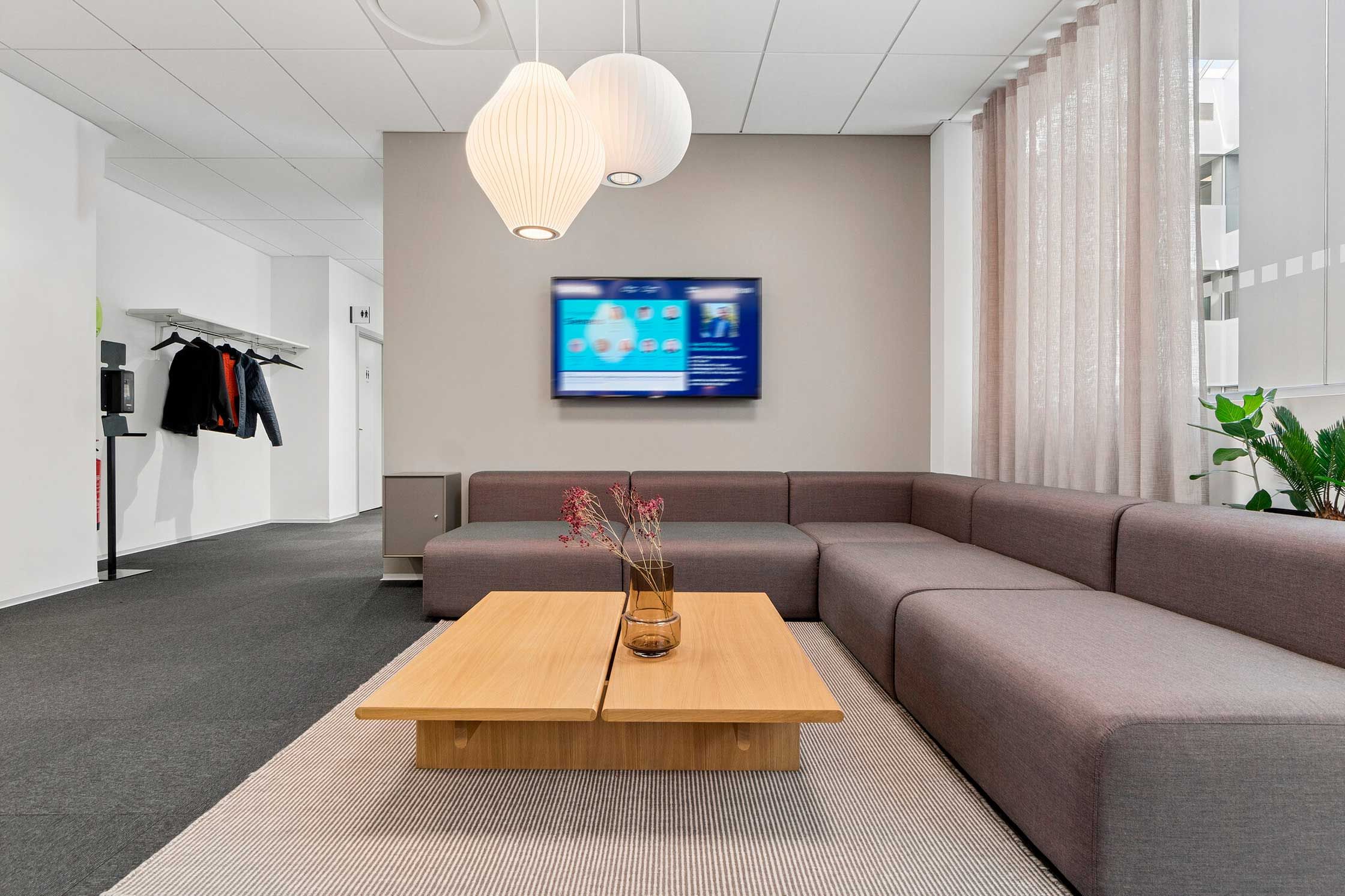 Kontorlokaler med moderne møbler og tv på væggen. Fællesområde med sofa, træbord og plante. Hængelys og tøjreoler.