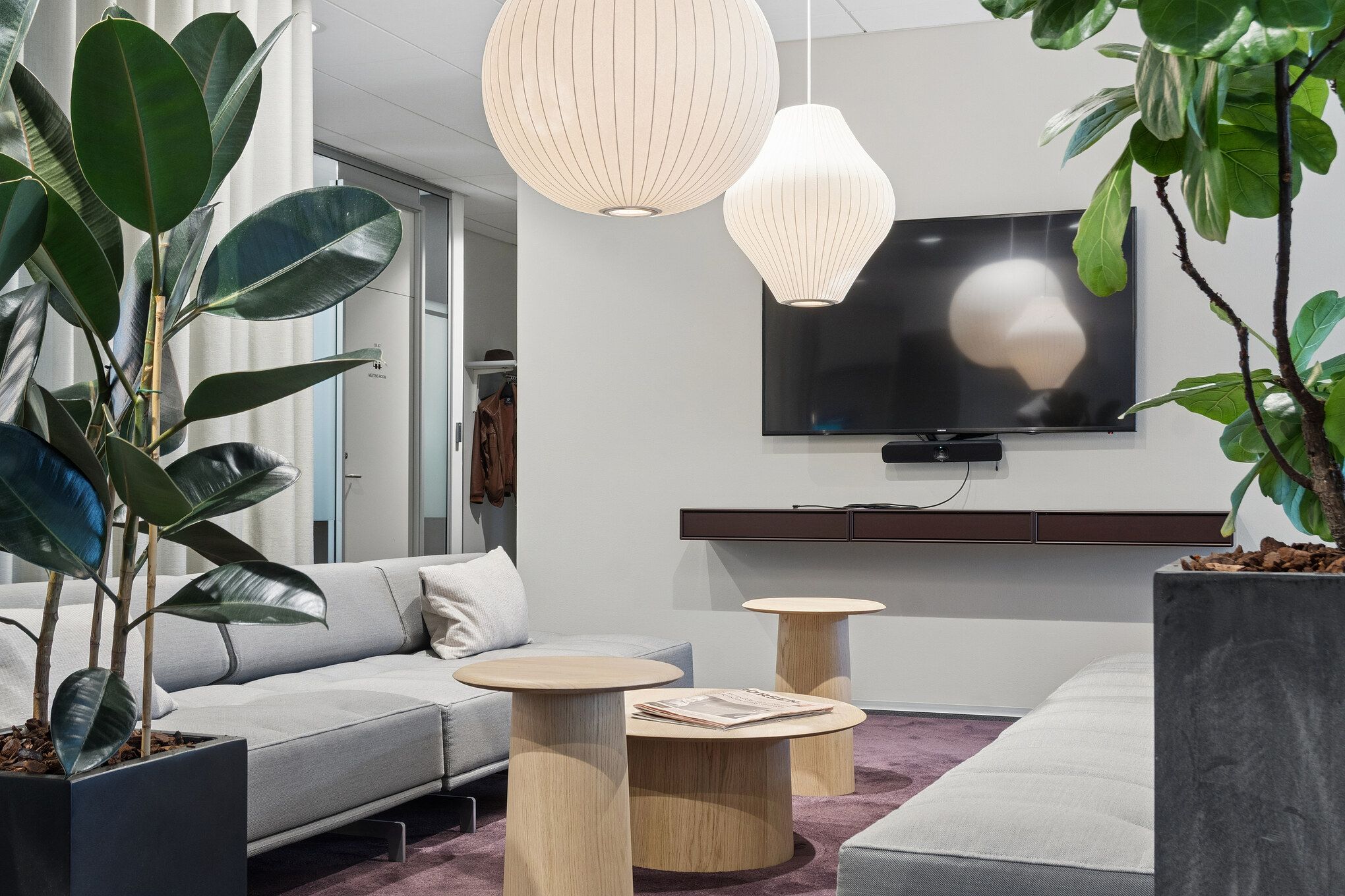 Kontorlokaler med moderne sofa, træborde, lyse lampekroner, tv og store planter i store potter og lyse vægge
