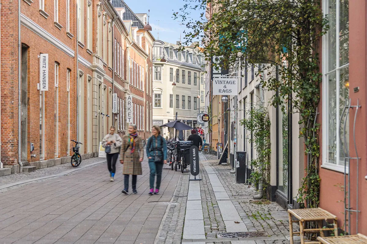 To kvinder går ad en gågade med butikker og gamle bygninger, der er dækket af grønne planter, og en vintage og tæpper-skilt er hængende på en bygning.
