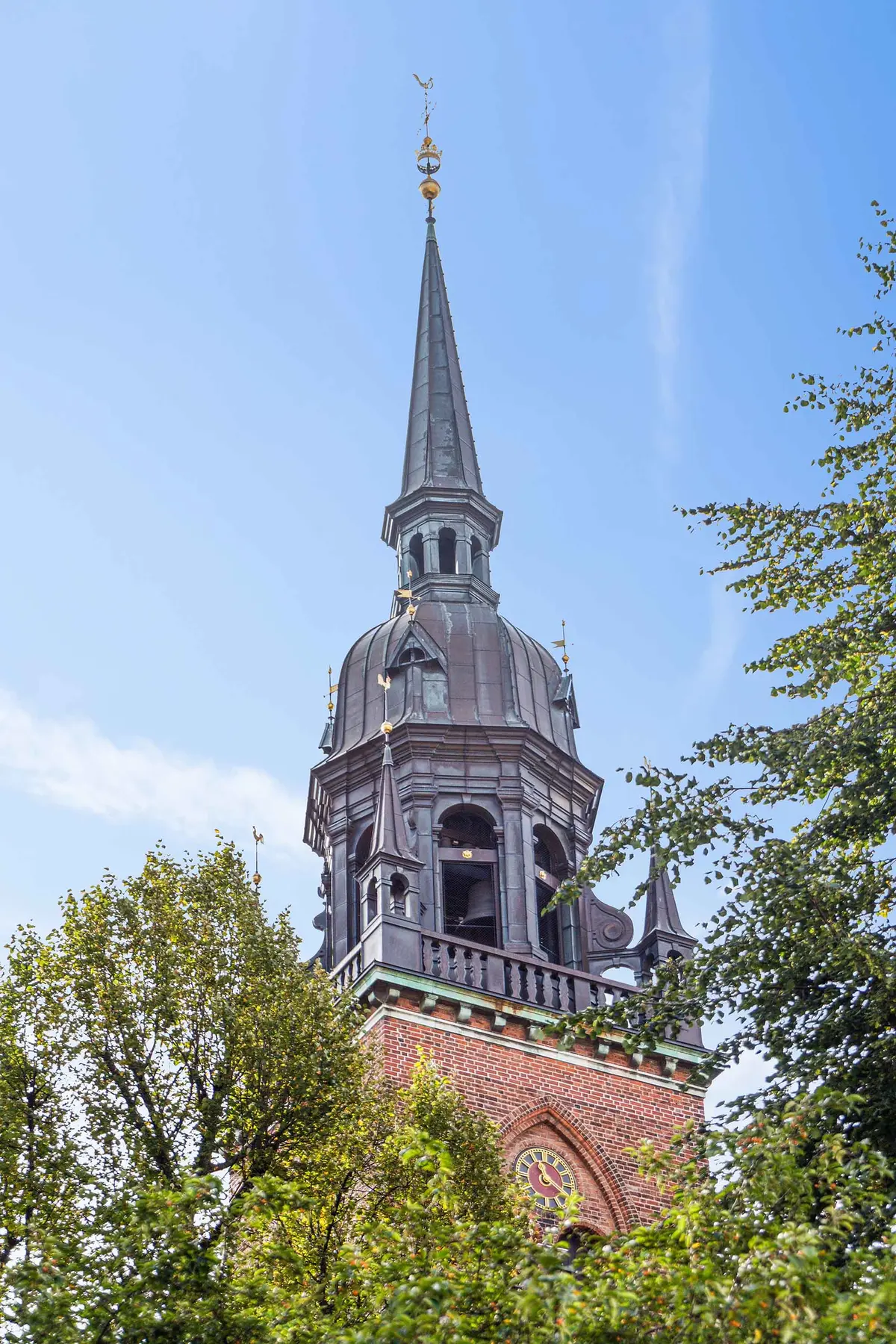 En kirke med spir og klokketårn, udsmykket med ur og vindrose, omgivet af grønne træer og solrig himmel