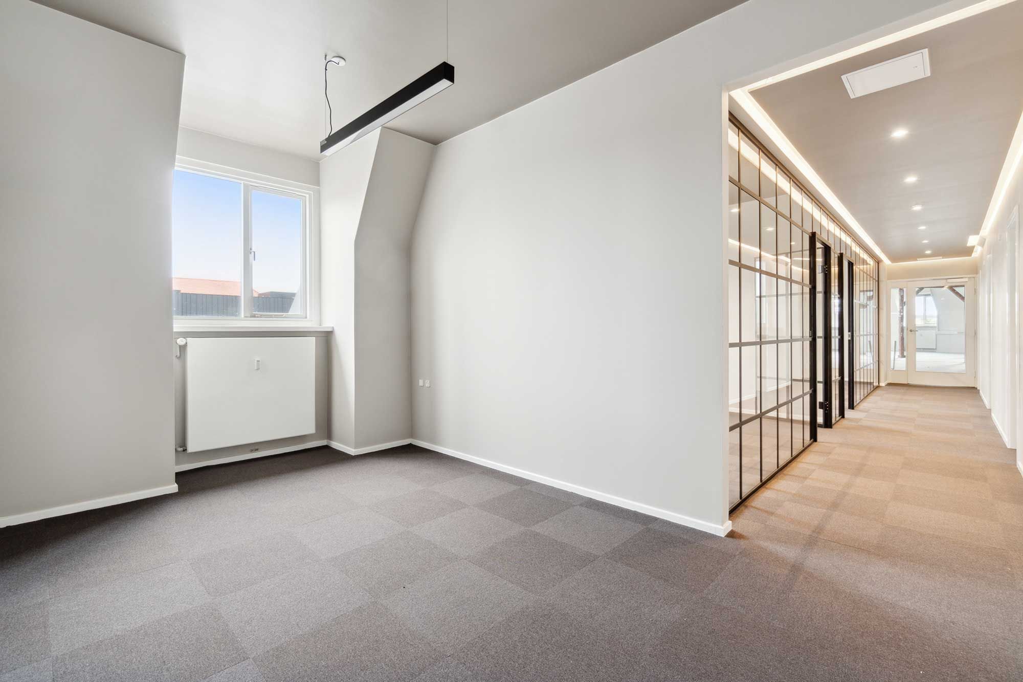 Tomme kontorrum med glasdøre, lysekroner, hvide vægge, gulvbelægning og et vindue med udsigt til en bygning og rødt tag