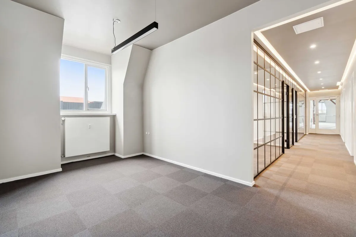 Tomme kontorrum med glasdøre, lysekroner, hvide vægge, gulvbelægning og et vindue med udsigt til en bygning og rødt tag