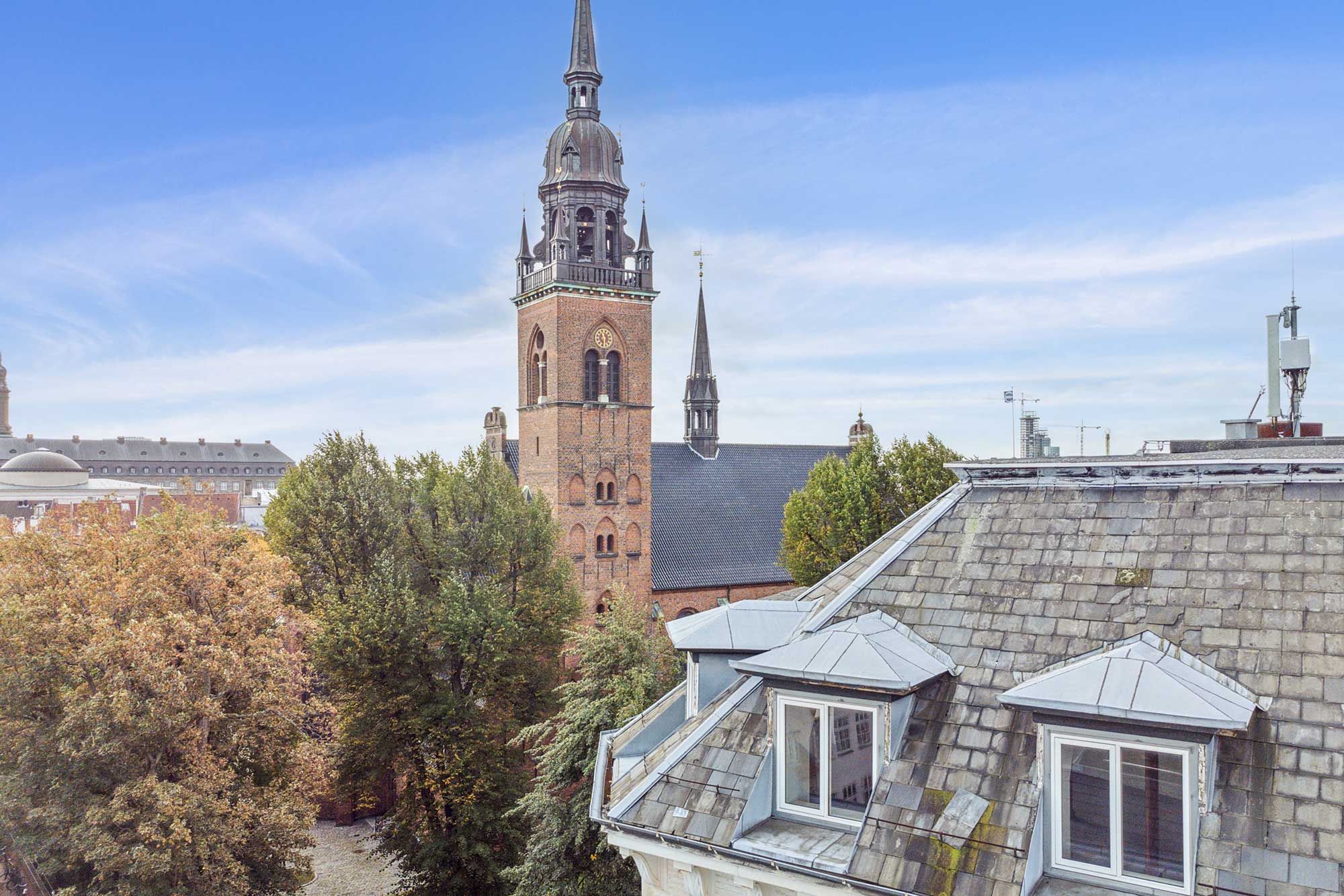 Historisk bygning med spir, kirke og tårn i baggrunden. Lysblå himmel, røde og gule blade, lyse vindueskarm og røde tagplader