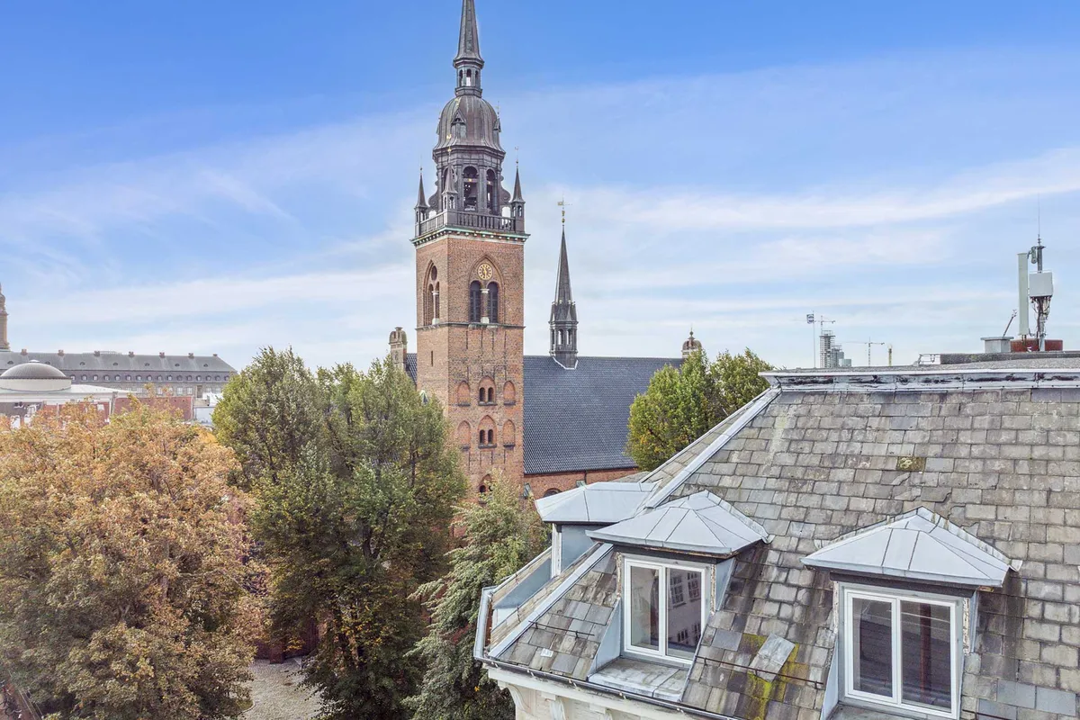 Historisk bygning med spir, kirke og tårn i baggrunden. Lysblå himmel, røde og gule blade, lyse vindueskarm og røde tagplader