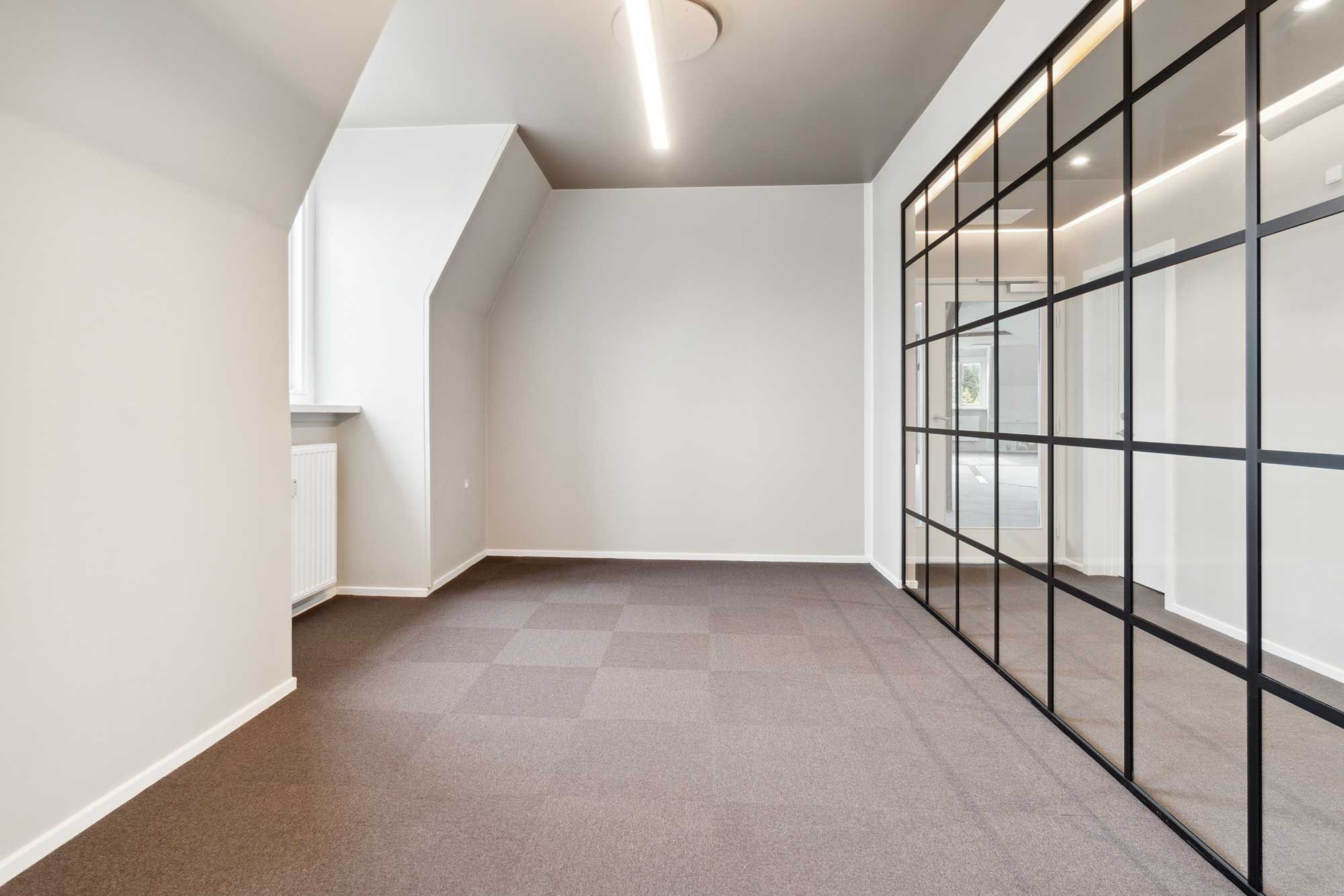 En stor og rummelig kontorlokal i hjertet af København K med udsigt og højloft. Væggene er hvide, gulvet er i lysegråt og der er en glasdør med mørke glas.