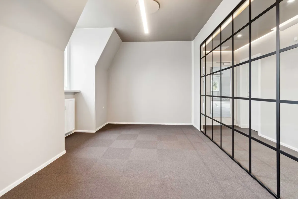 En stor og rummelig kontorlokal i hjertet af København K med udsigt og højloft. Væggene er hvide, gulvet er i lysegråt og der er en glasdør med mørke glas.