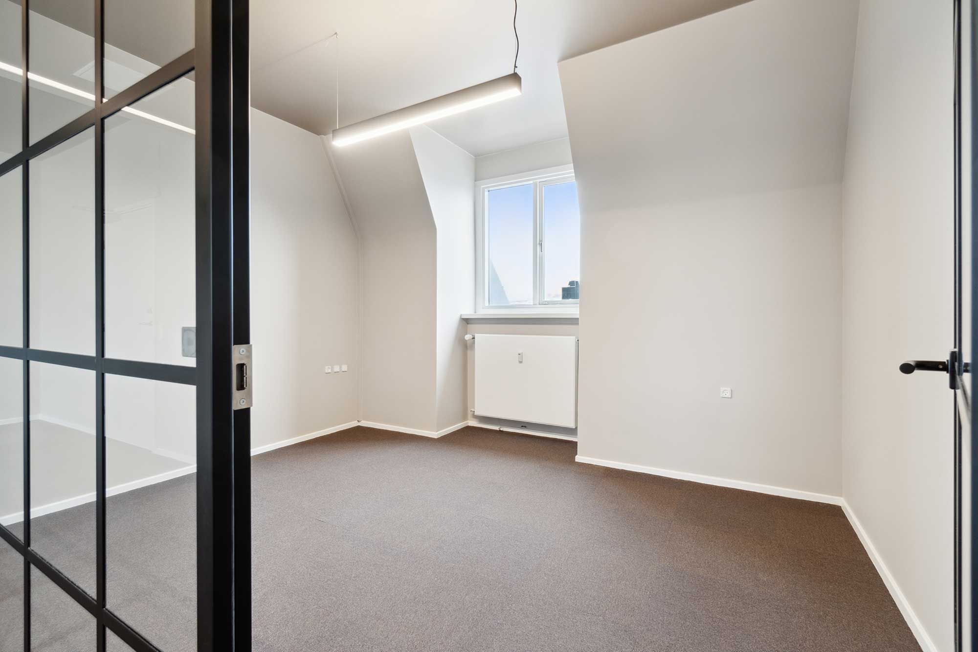 En tom kontorlokal med hvide vægge og gulv, hvor der er en vinduesåbning med en hvid vindueskarm og et hængende lys. Loftet er hvidt og der er et radiator.