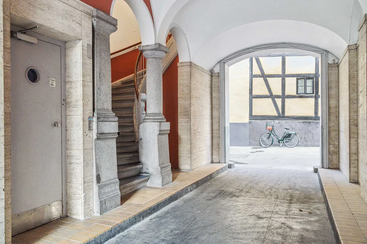 Indenfor en gammel bygning er der en hal med to søjler, en trappe og en dør til højre. På højre side er der et vindue med en cykel udenfor.