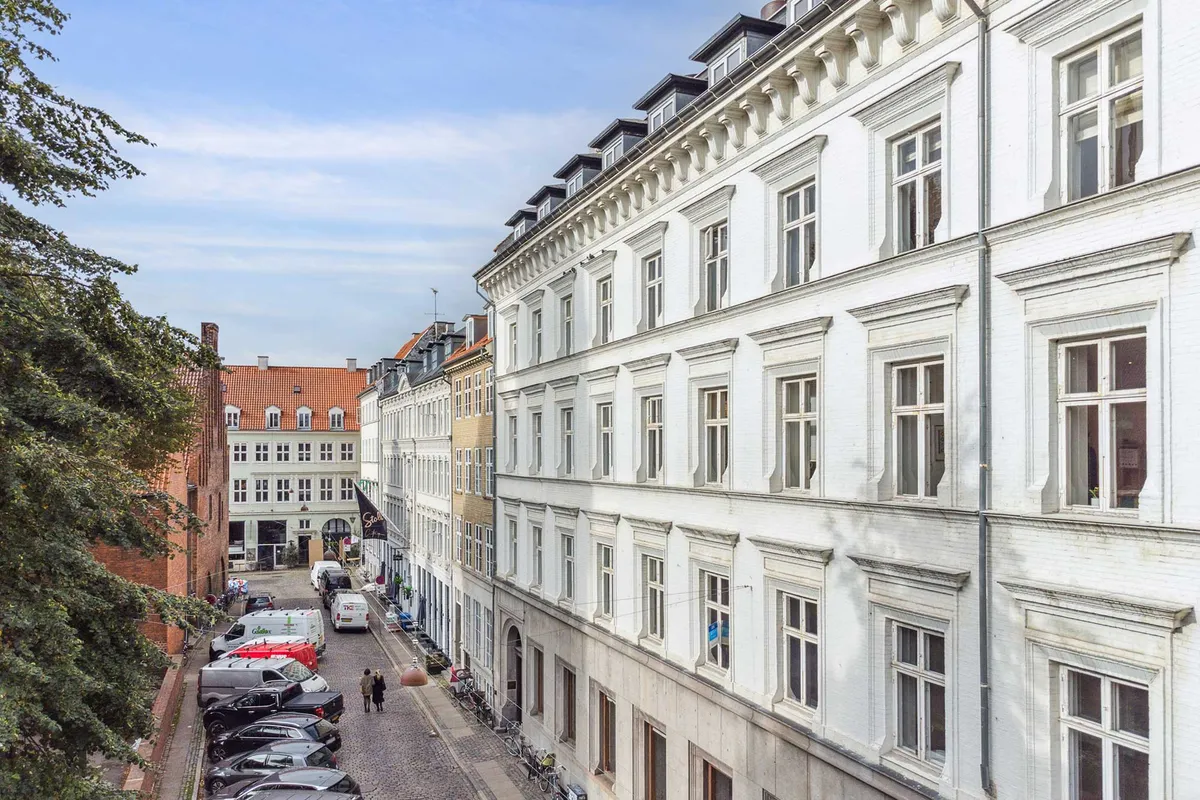 Kontor i charmerende, klassisk bygning med udsigt over København, parkerede biler og enkeltpersoner, høje huse, vinduerne med lysegardiner og hvide vægge