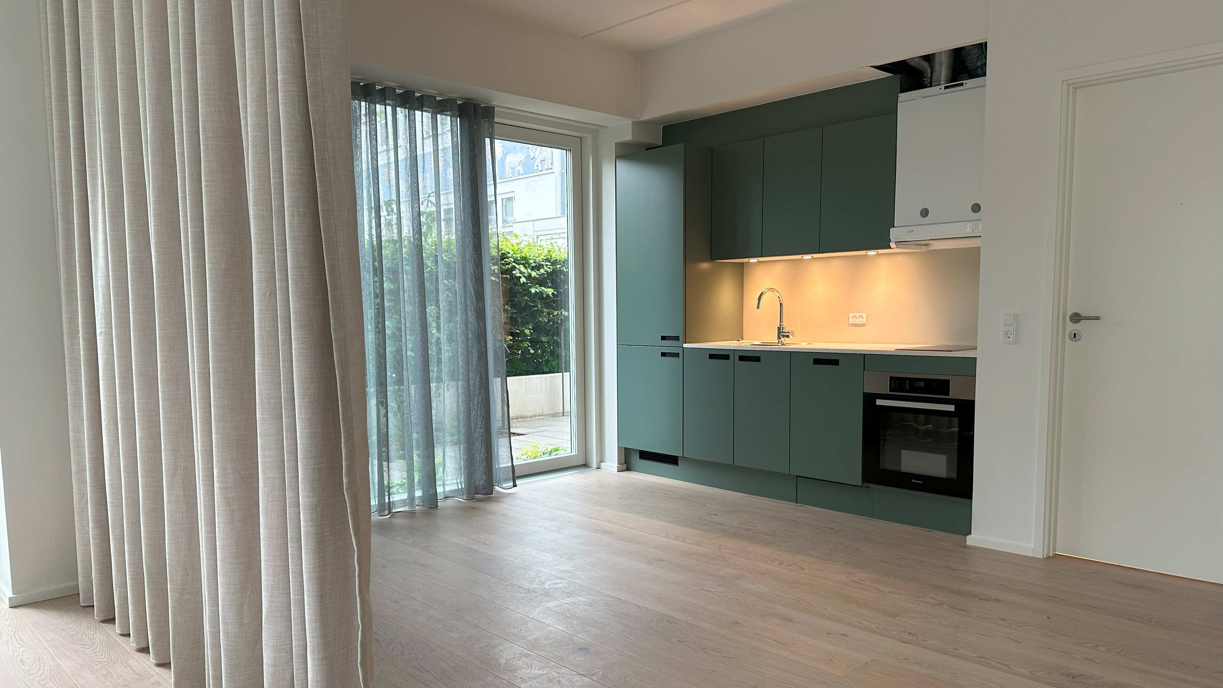 Lys stue med lysegrå gardiner og møbleret køkken med grønne skabe, moderne hvid indretning og trægulv