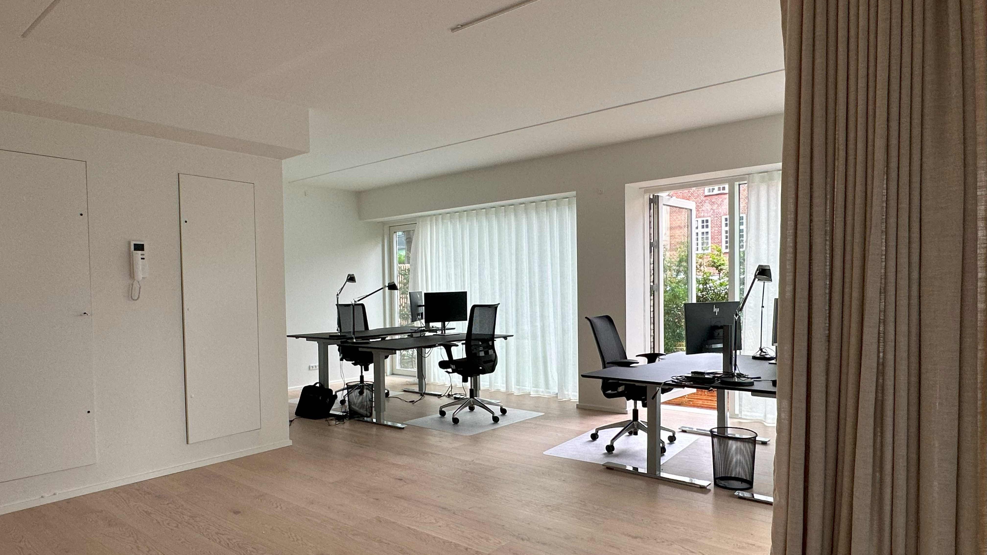 To kontorborde med to stole og monitorer, lukket gardin og skrivebordslampe samt en skraldespand.