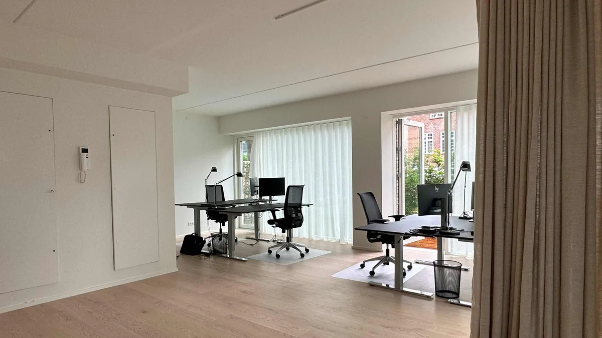 To kontorborde med to stole og monitorer, lukket gardin og skrivebordslampe samt en skraldespand.