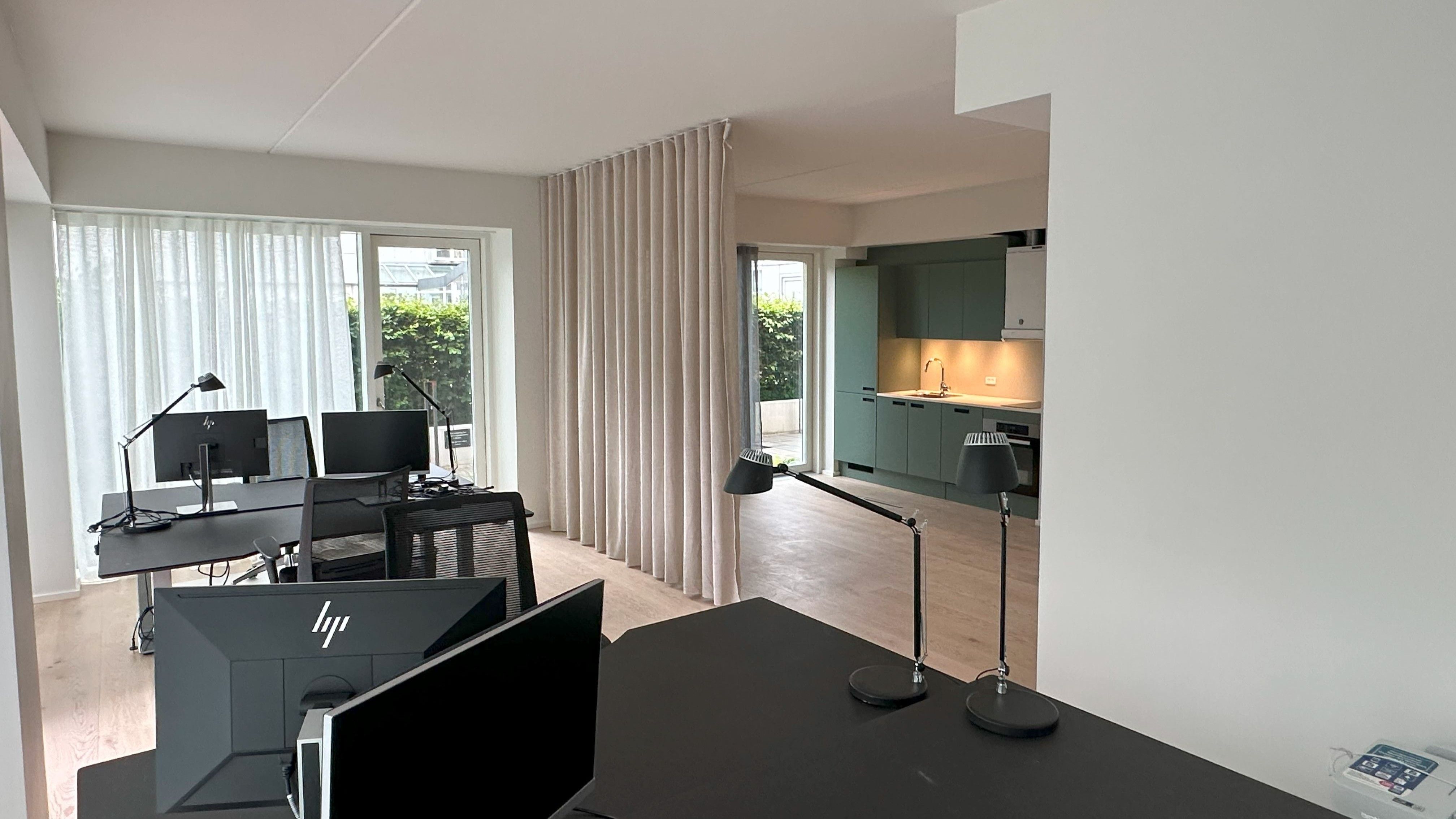 Et lyse og moderne kontor med en skrivebord og flere computere. Lamper og møbler er placeret rundt i rummet. En delvist åben køkkendel med hætte er synlig i baggrunden.