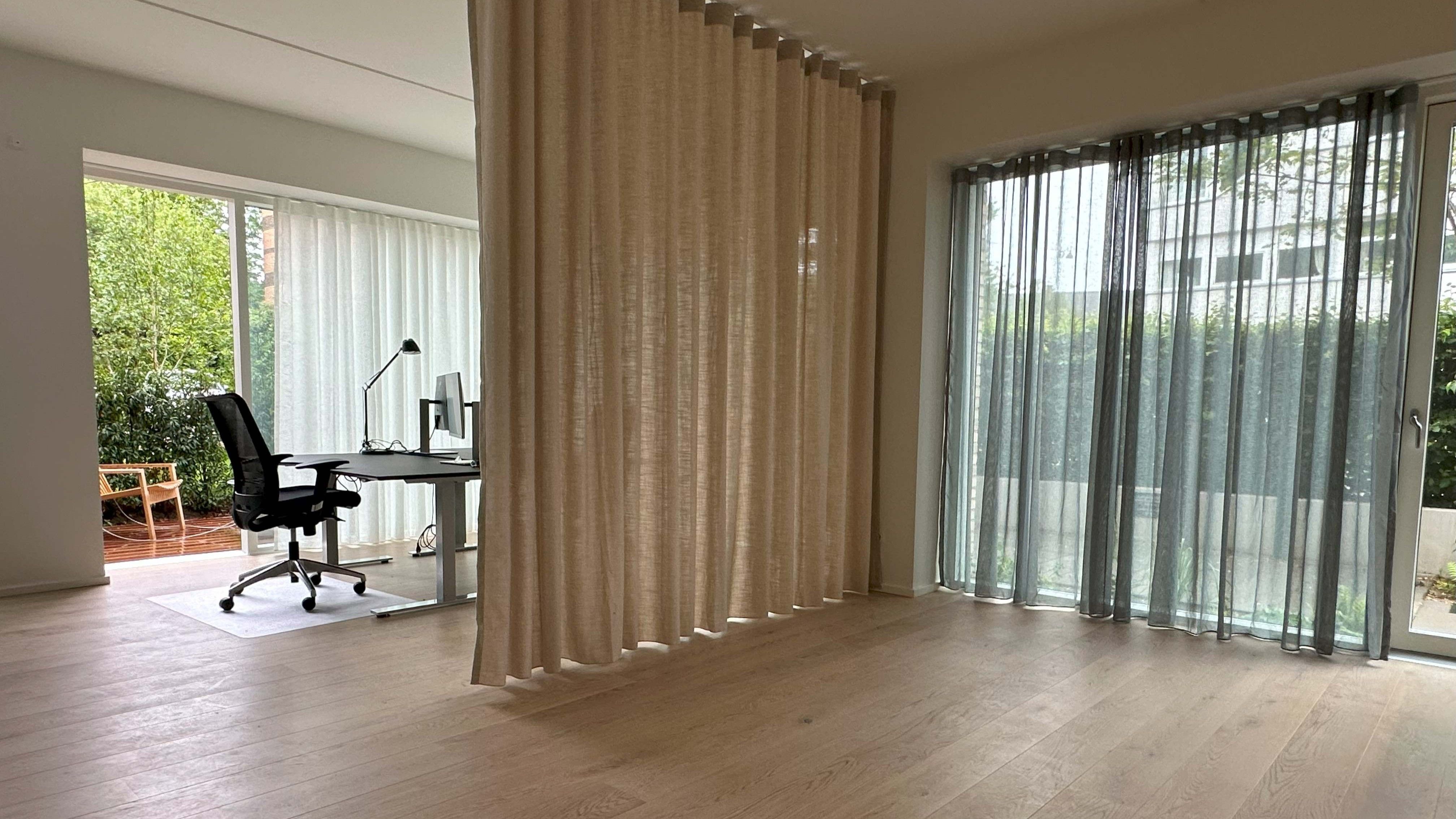 Foto af et moderne kontor med lyse gulvbelægning, højt loft og en stor skrivebord med computer, stol og lampe. Væggen er dækket af en beige gardin, der adskiller arbejdsområdet fra vinduespartiet