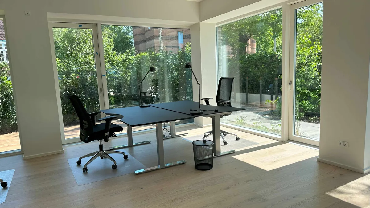 Kontorrum med to skriveborde og to stolpe til at sidde på. Et skrivebord med to skraldespande og et papirkurv på gulvet. To stolpe med hjul og en lampe.