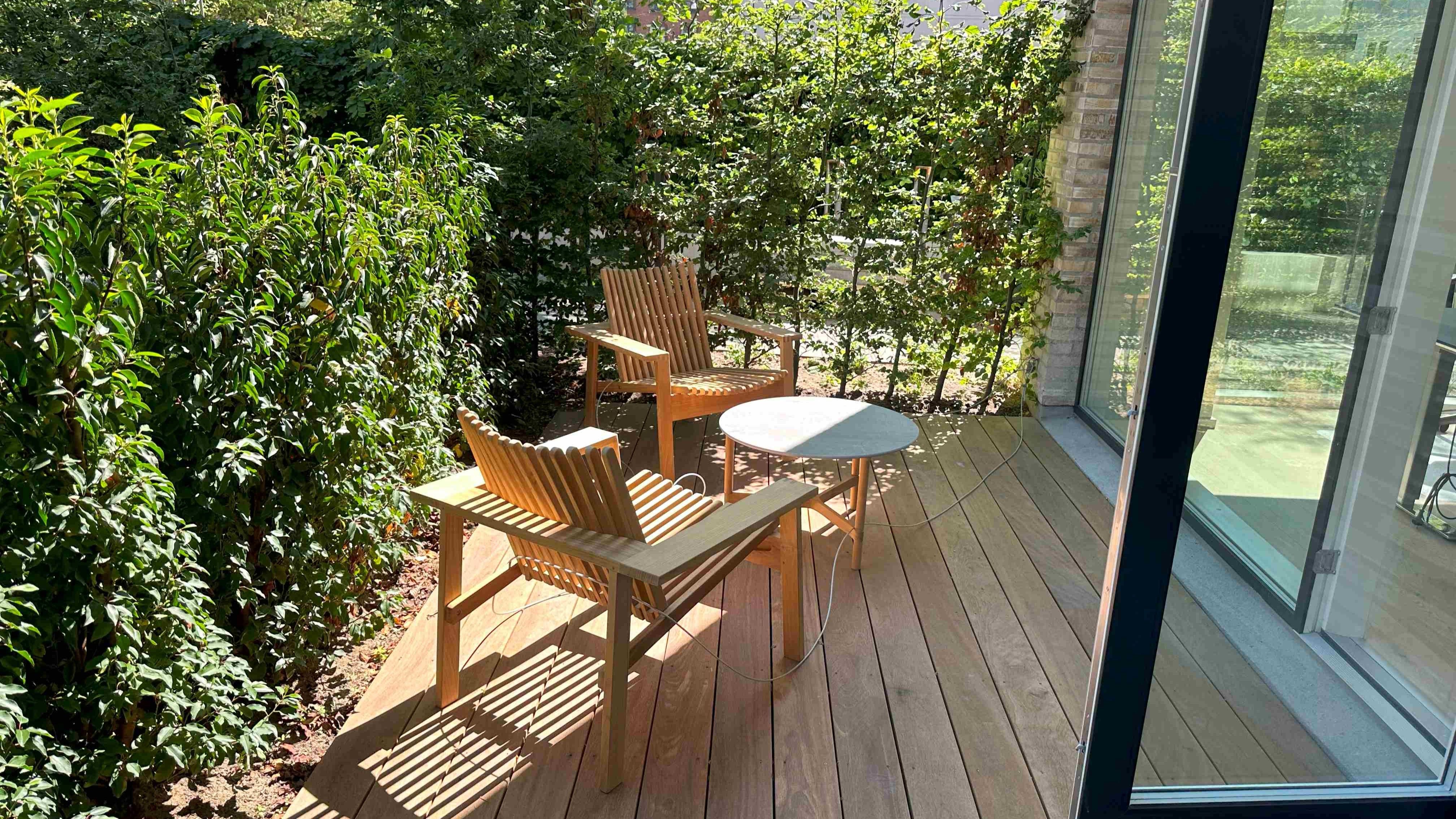 Et lille, træbelægget terrasseområde med to træstole og et rundt bord og udsigt til skov og grønne planter