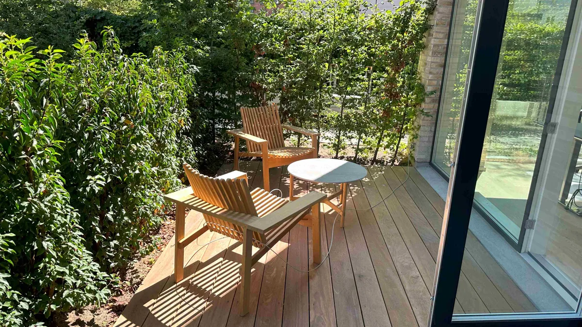 Et lille, træbelægget terrasseområde med to træstole og et rundt bord og udsigt til skov og grønne planter