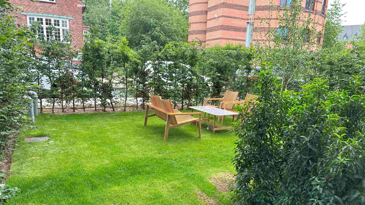 En lille have med lyse grønne græs, to træstole, et bord og høje grønne buske med buskplanter i baggrunden