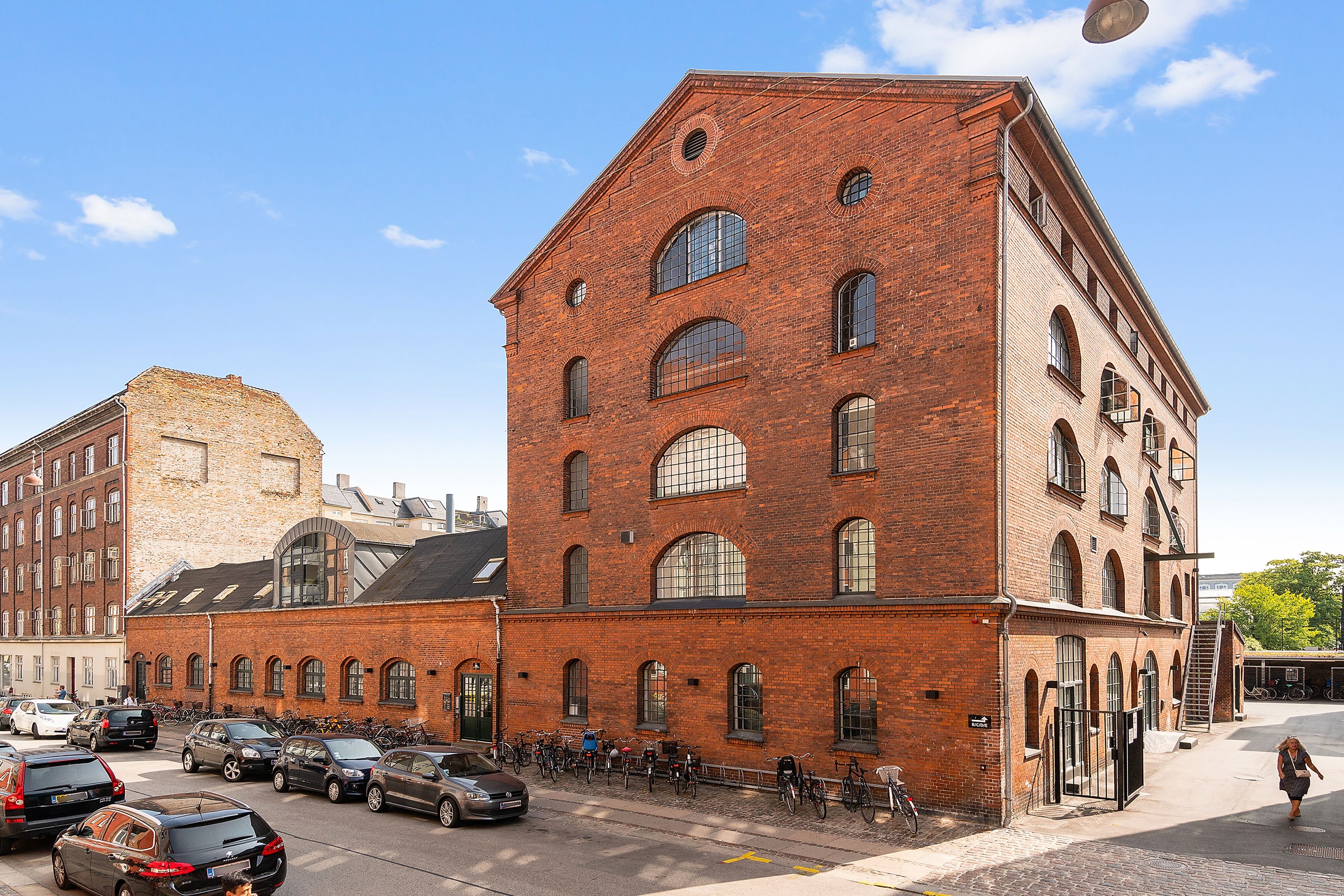 Mange cykler parkeret udenfor kontorer på Viborggade 70. Kvinde går på gågade med kørefelt. Moderne kontorer med røde mursten og lyse vinduer. Solskin og lyse himmel med skyer.