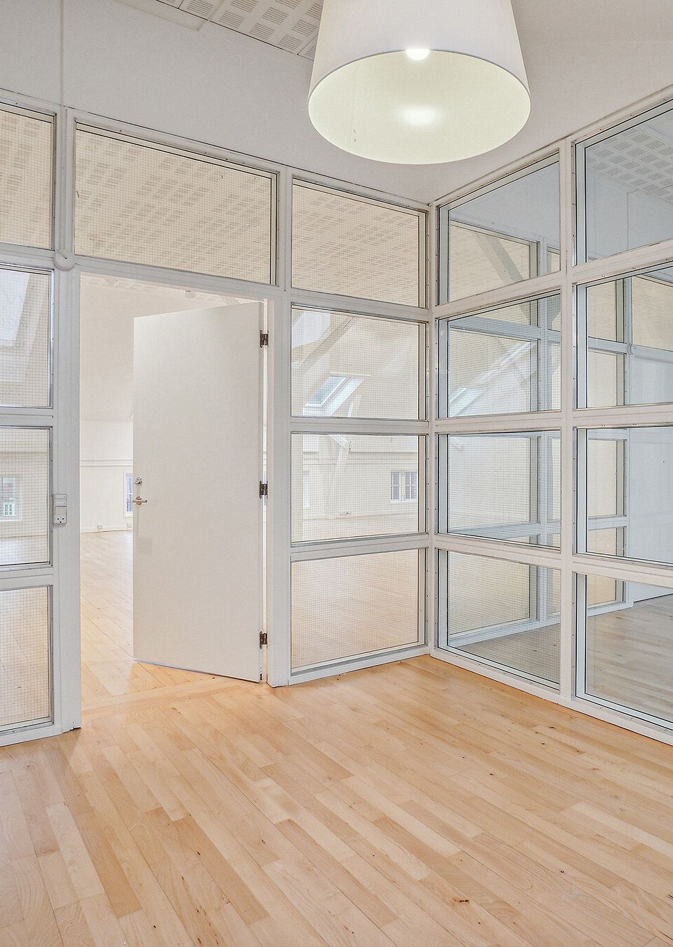En højloftet kontorbygning med lyse, glasvinduer og lysekroner og åbne døre og trægulve. Lyst og moderne kontormiljø med store vinduer og højloftet.