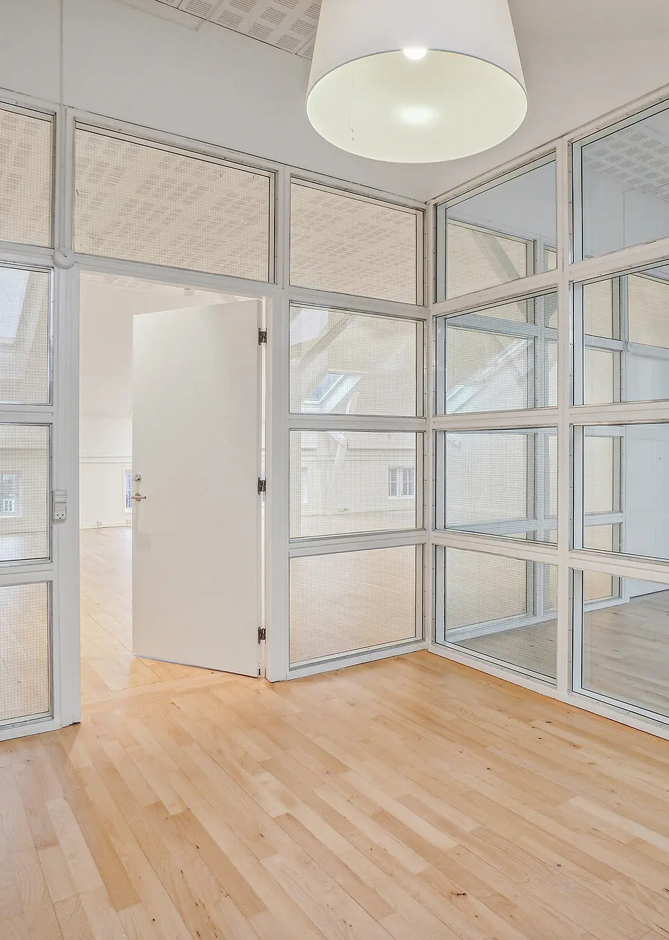 En højloftet kontorbygning med lyse, glasvinduer og lysekroner og åbne døre og trægulve. Lyst og moderne kontormiljø med store vinduer og højloftet.