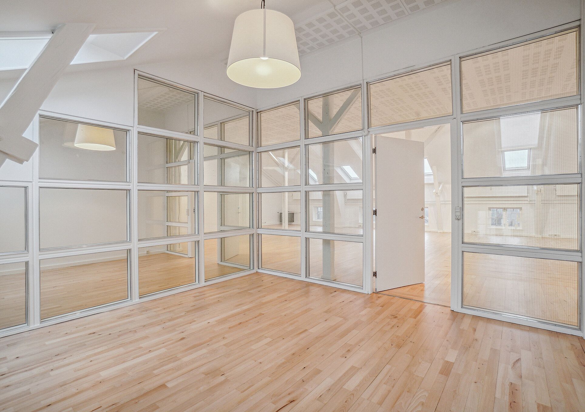 En højloftet kontorlokal med glasvægge og massivt trægulv i tidligere fabriksbygning. Lyst og rummeligt kontormiljø med lysekroner og lyse lofter.