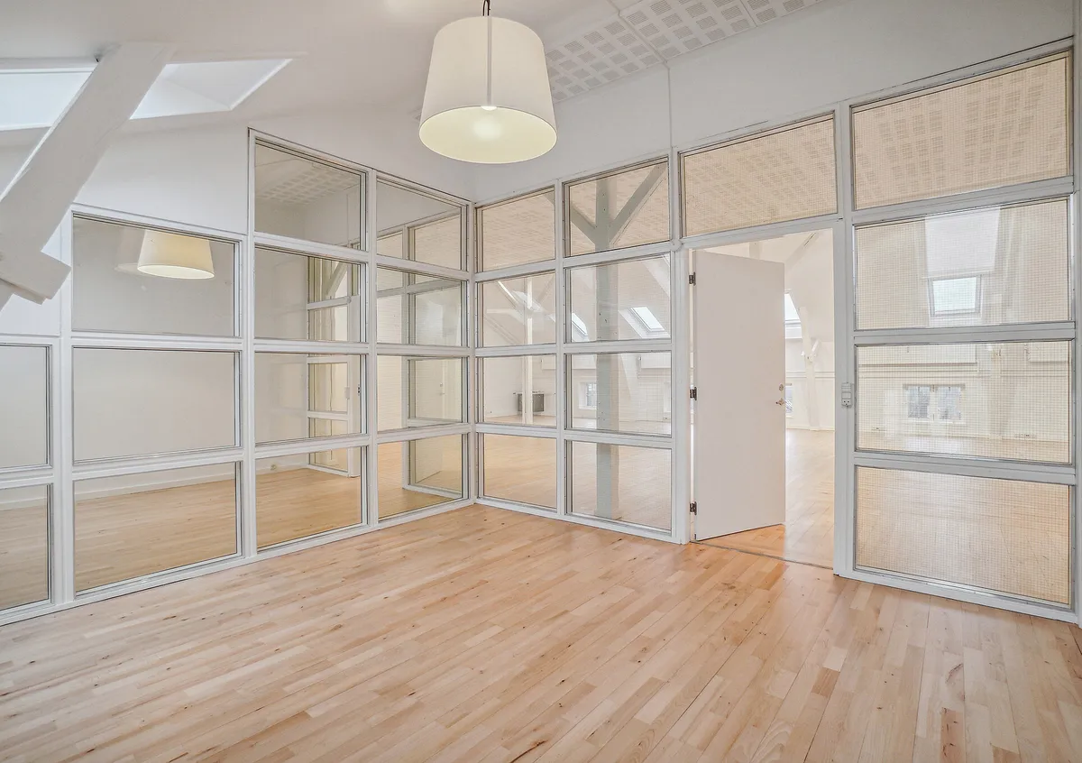 En højloftet kontorlokal med glasvægge og massivt trægulv i tidligere fabriksbygning. Lyst og rummeligt kontormiljø med lysekroner og lyse lofter.