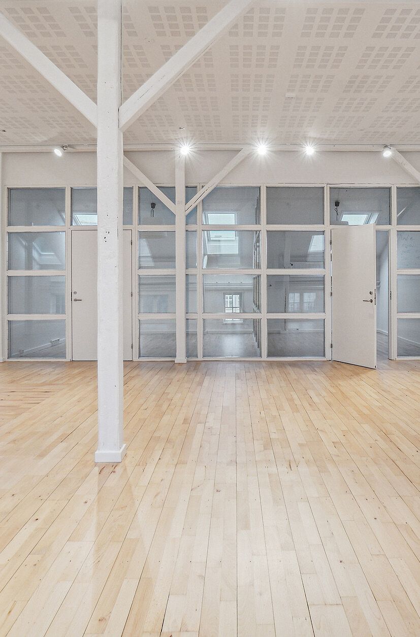 En højloftet kontorlokal i tidligere fabriksbygning med lyse gulvplanker, hvide vægge, store glasvinduer og en stor, åben dør.