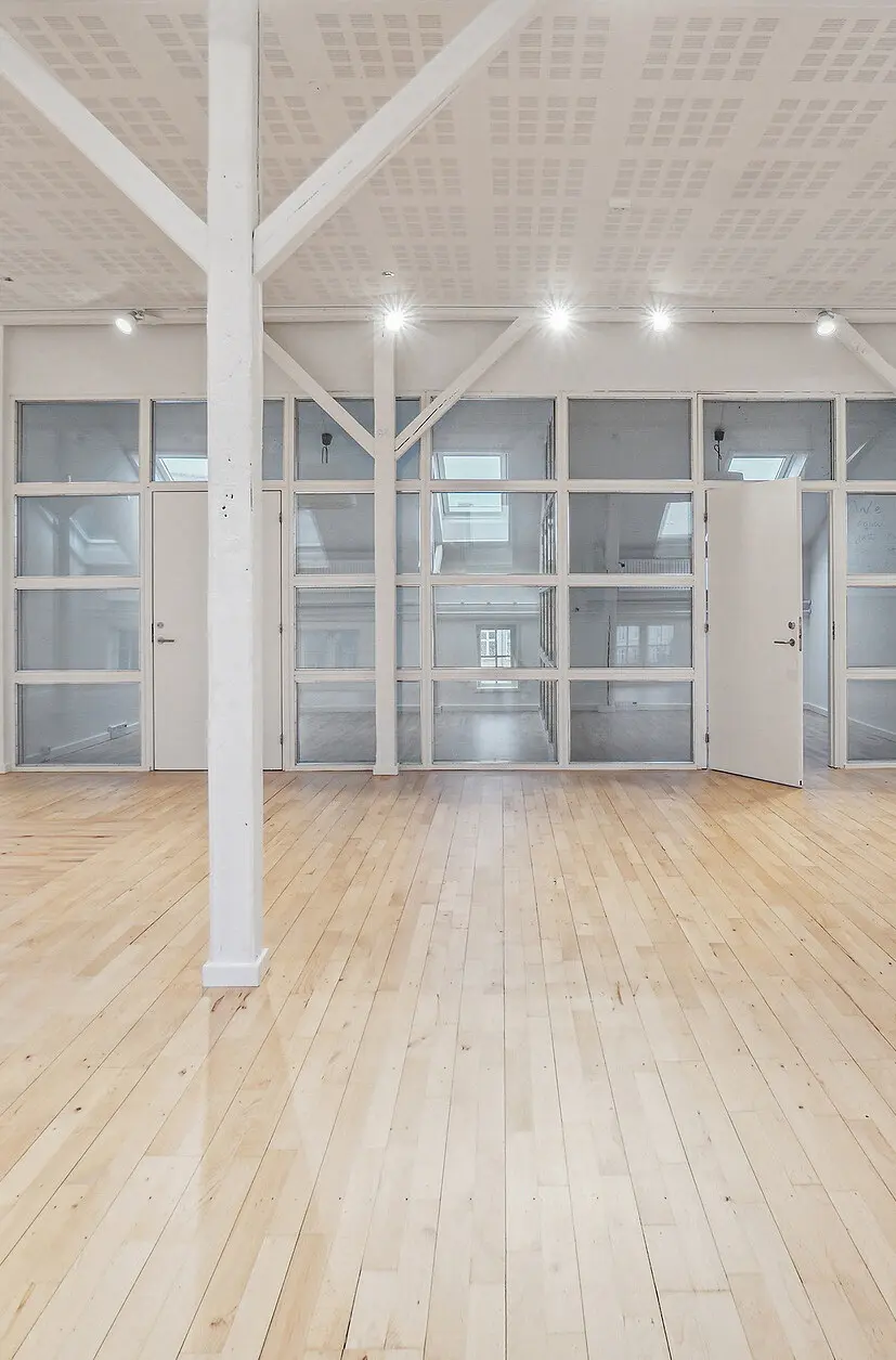 En højloftet kontorlokal i tidligere fabriksbygning med lyse gulvplanker, hvide vægge, store glasvinduer og en stor, åben dør.