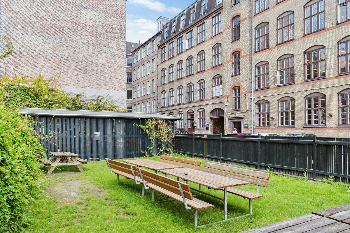 En stor grøn græsplæne med parkborde og bænke i et industrielt miljø med gamle bygninger og høje gavle.