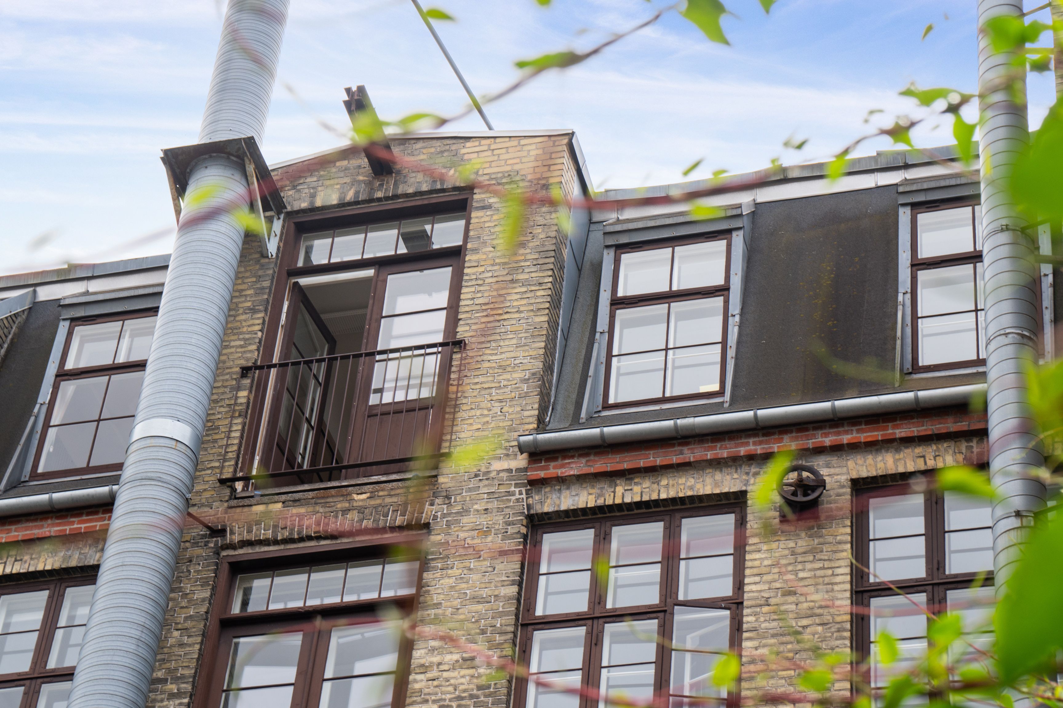 Flot kontor med store vinduer, røde mursten og industrielle røgkammere. Balkon og vinduer med brune rammer og lysegardiner.