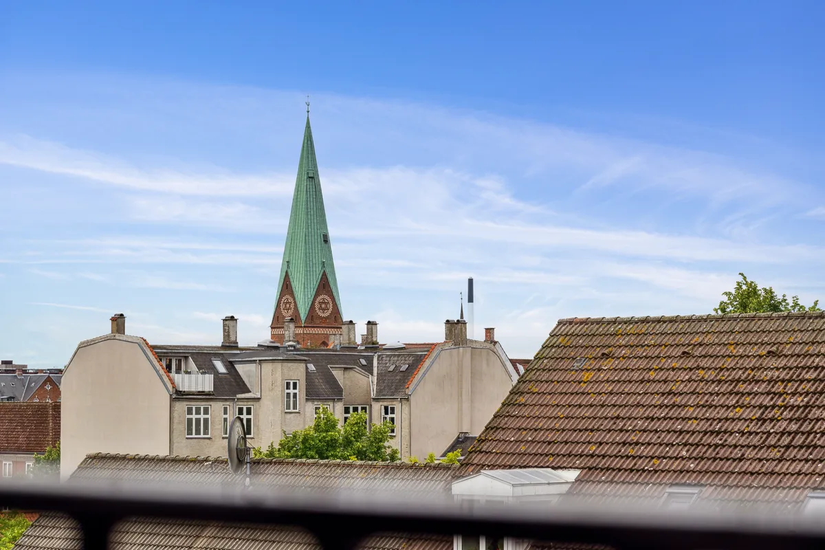 Byudsigt med grønt spir og kirkeklokke på Nørrebrogade 45D, 4. th, 2200 København N. Blik mod lyserøde tag og søjler. Solskin og lyseblå himmel med skyer.