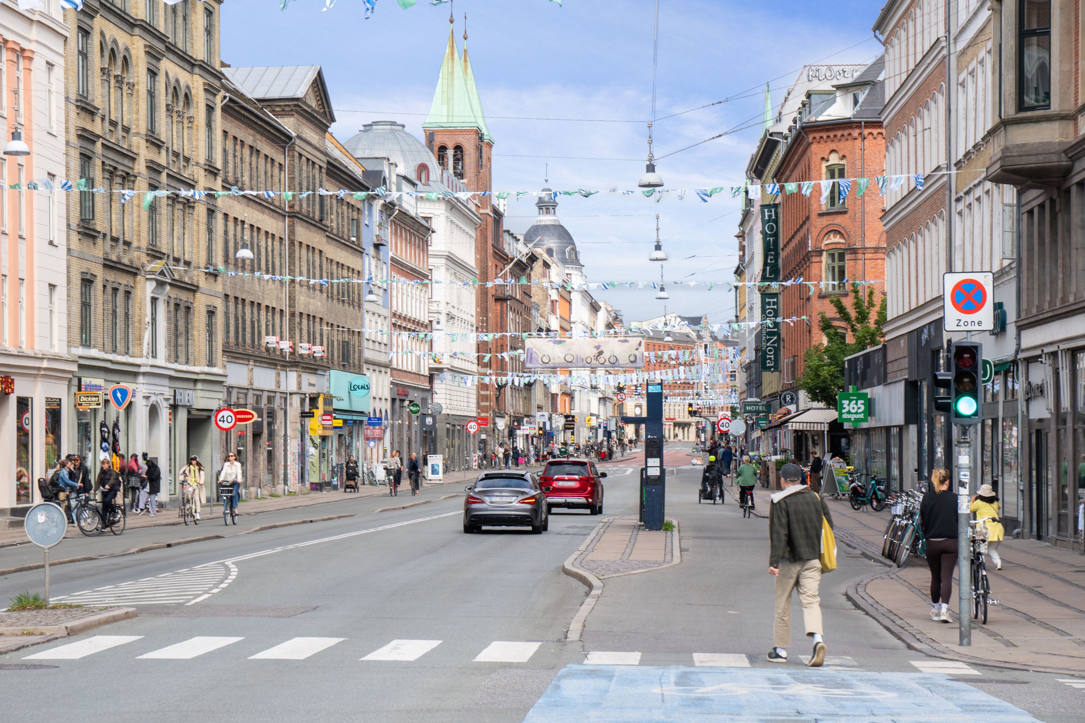 Mennesker går rundt på en gågade i København med biler og cyklister, og der er en stolthed i byens historie og bygninger