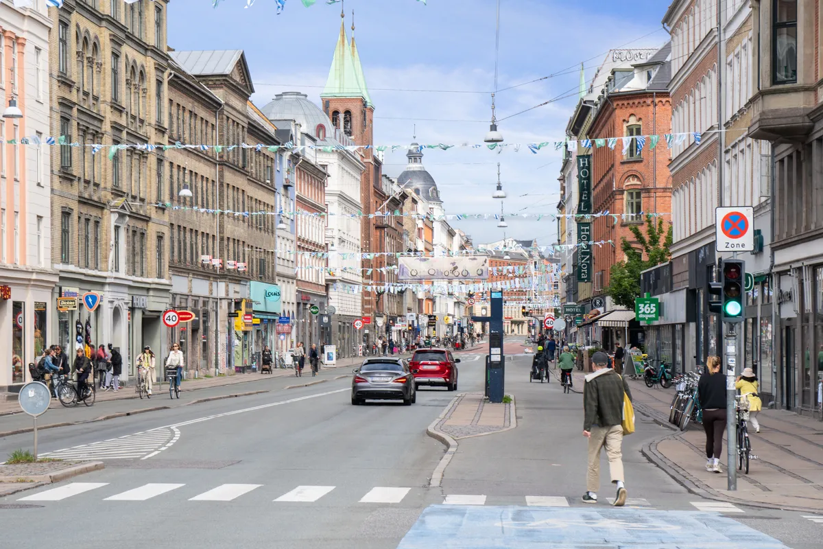 Mennesker går rundt på en gågade i København med biler og cyklister, og der er en stolthed i byens historie og bygninger