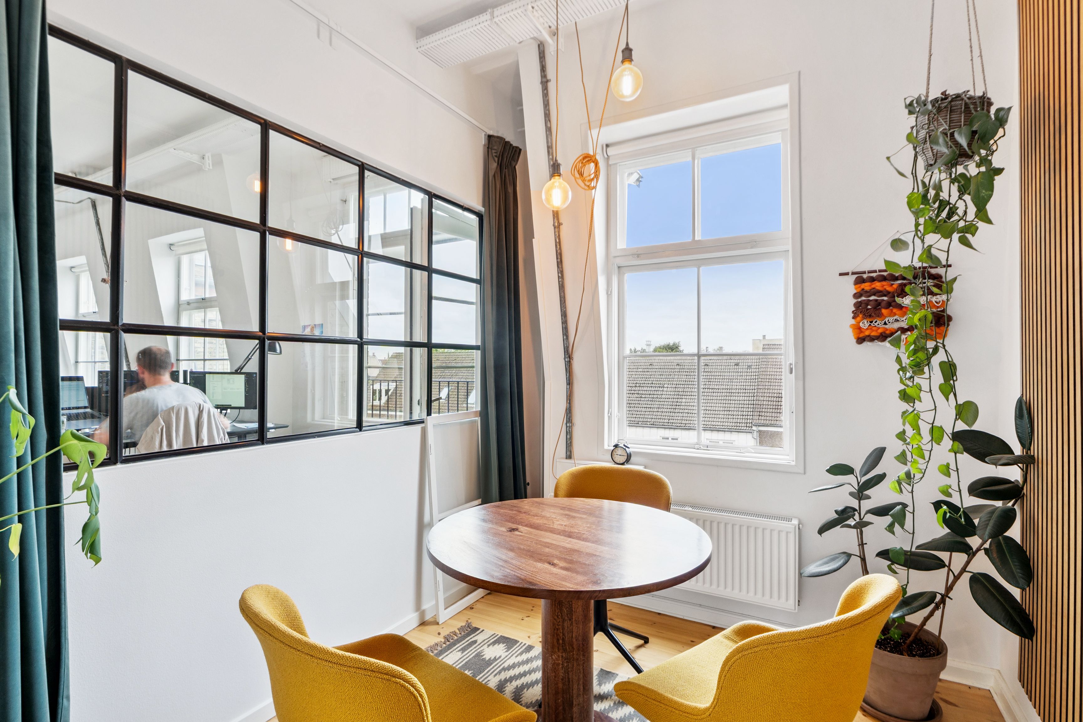 En flot kontorrum med et rundt bord og gule stole, plante, spejl og vinduer med udsigt til gaden. En mand sidder med en computer og arbejder i spejlet.