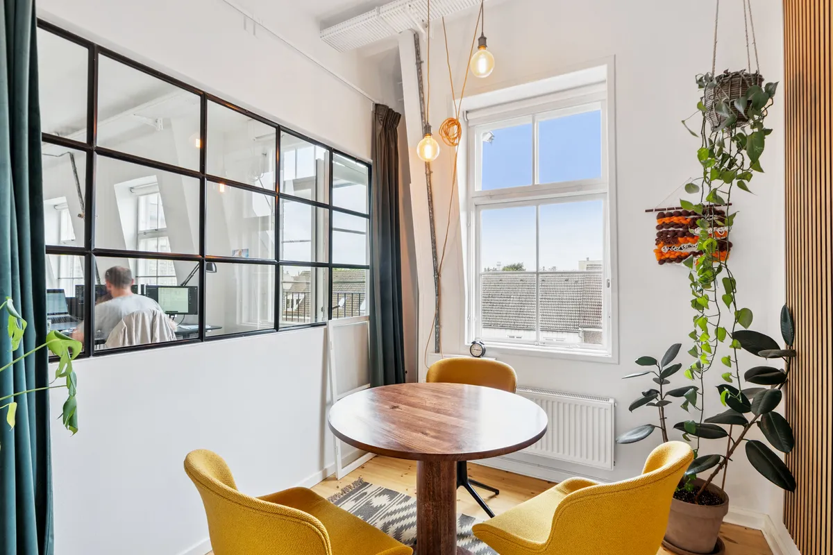 En flot kontorrum med et rundt bord og gule stole, plante, spejl og vinduer med udsigt til gaden. En mand sidder med en computer og arbejder i spejlet.