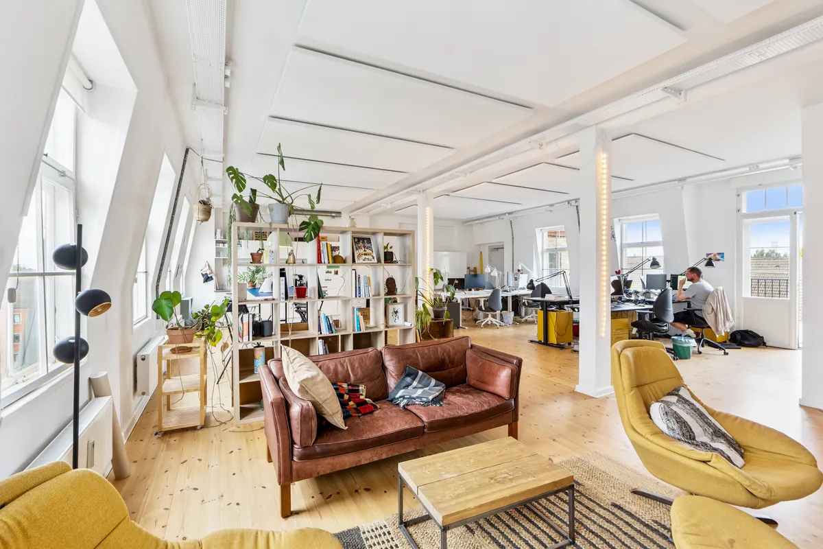 Moderne kontor med loftsrum og lyse lofter. Et mandsfigur sidder ved et skrivebord. Gulvet er lavet af træ og er lysebrunt. Et skrivebord og en sofa er placeret midt i rummet.