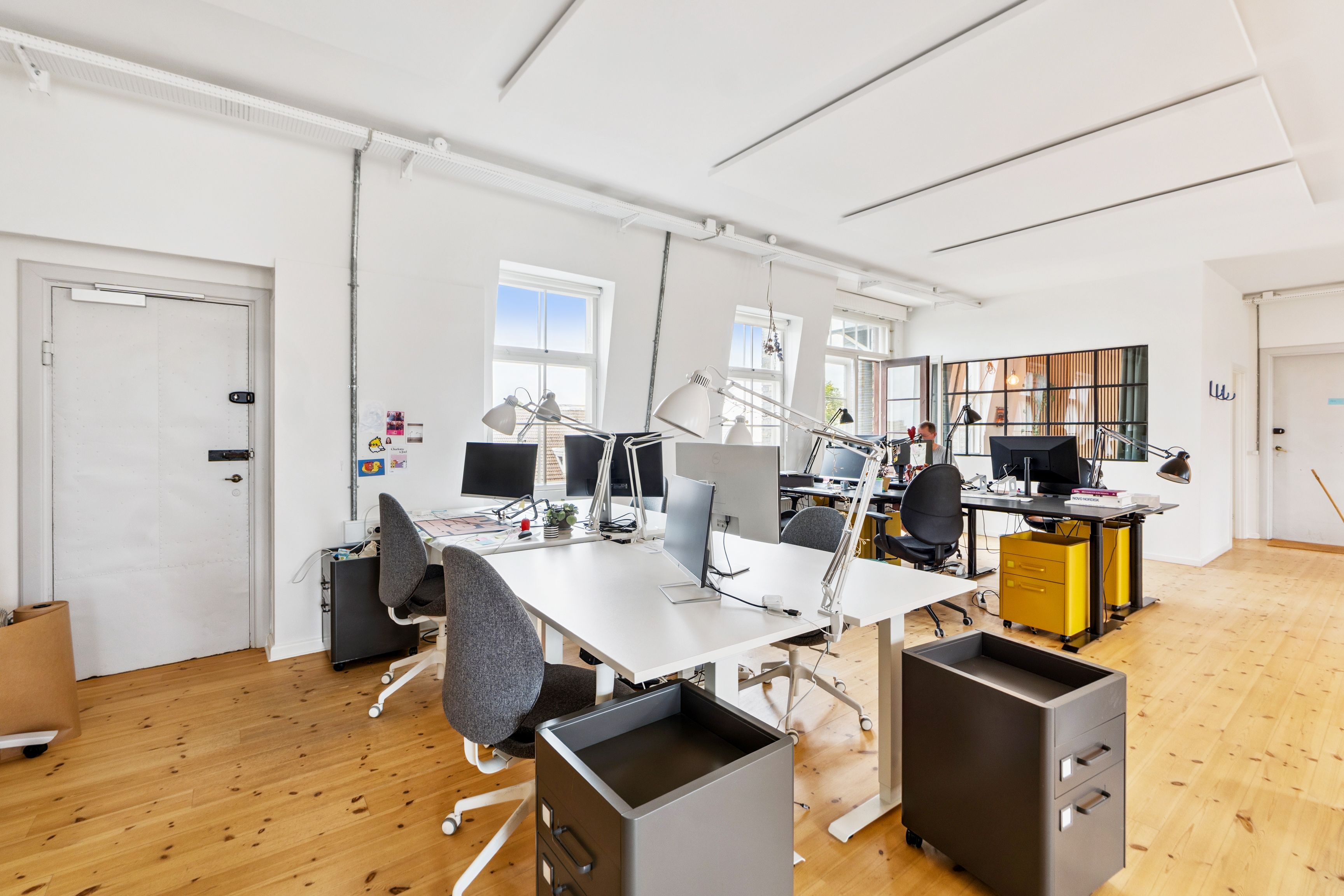 Flot kontor med højt til loftet og store vinduer. Værelset er fuldt møbleret med skriveborde, stole og computere. Der er også en person, der sidder og arbejder på skrivebordet.