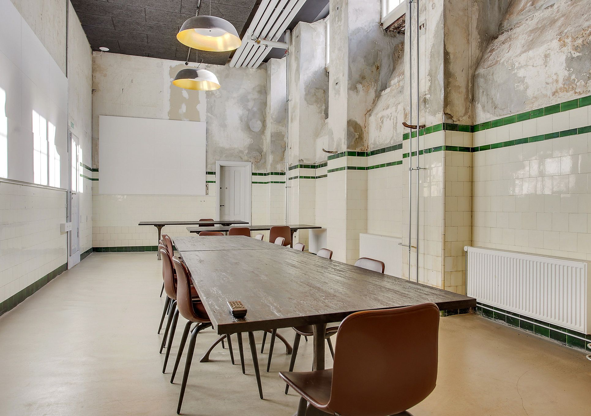 Flot kontor med lange borde og lyse loftslamper i en rummelig og industrielt indrettet stue med hvide og grønne vægfliser og radiatorer