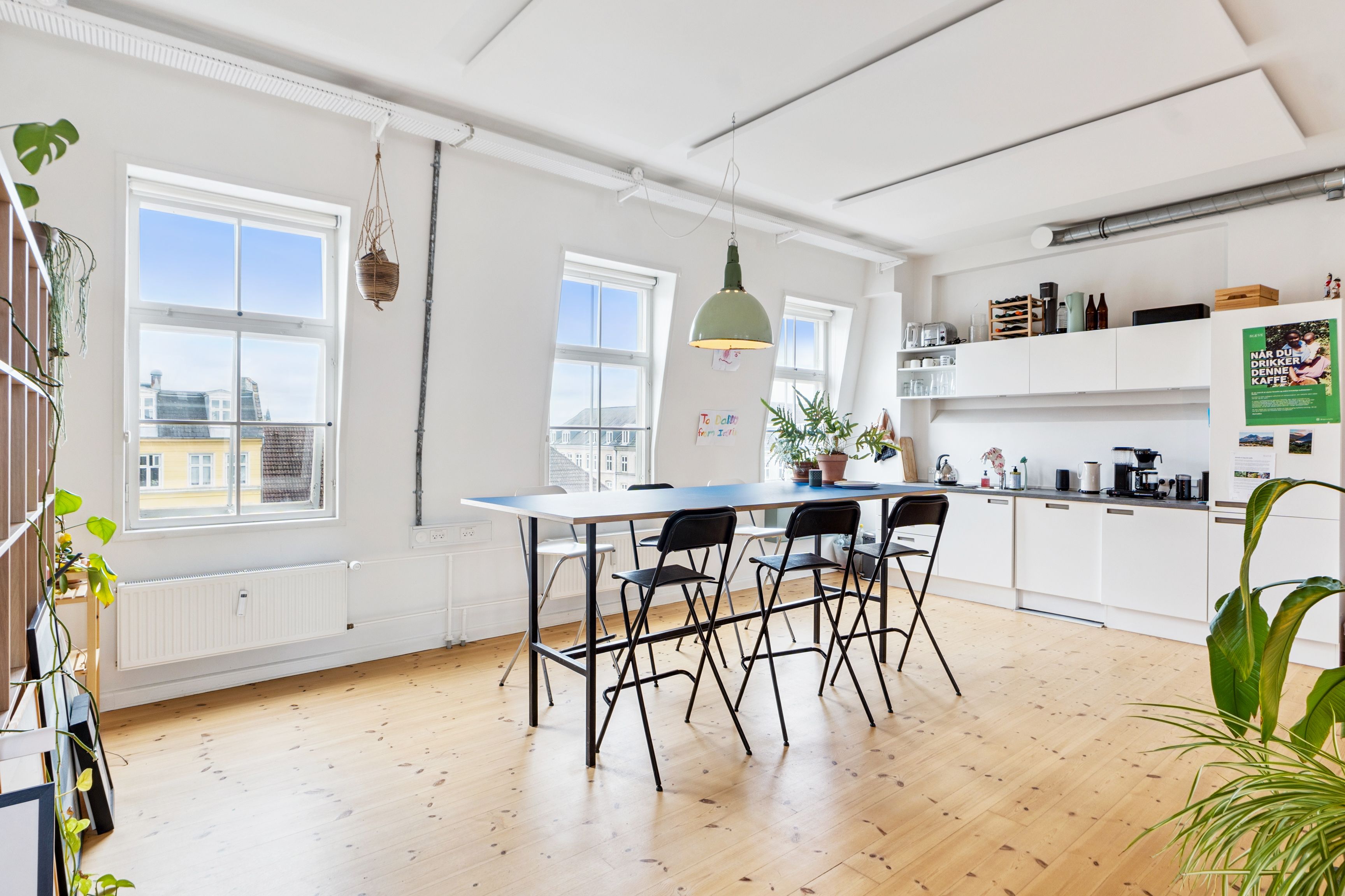 Flot kontor med højloft, lysevinduer og højloft. Bord med fire stole og plante. Køkken med moderne apparater og højloft. Loftsvinduer med udsigt til Nørrebro.