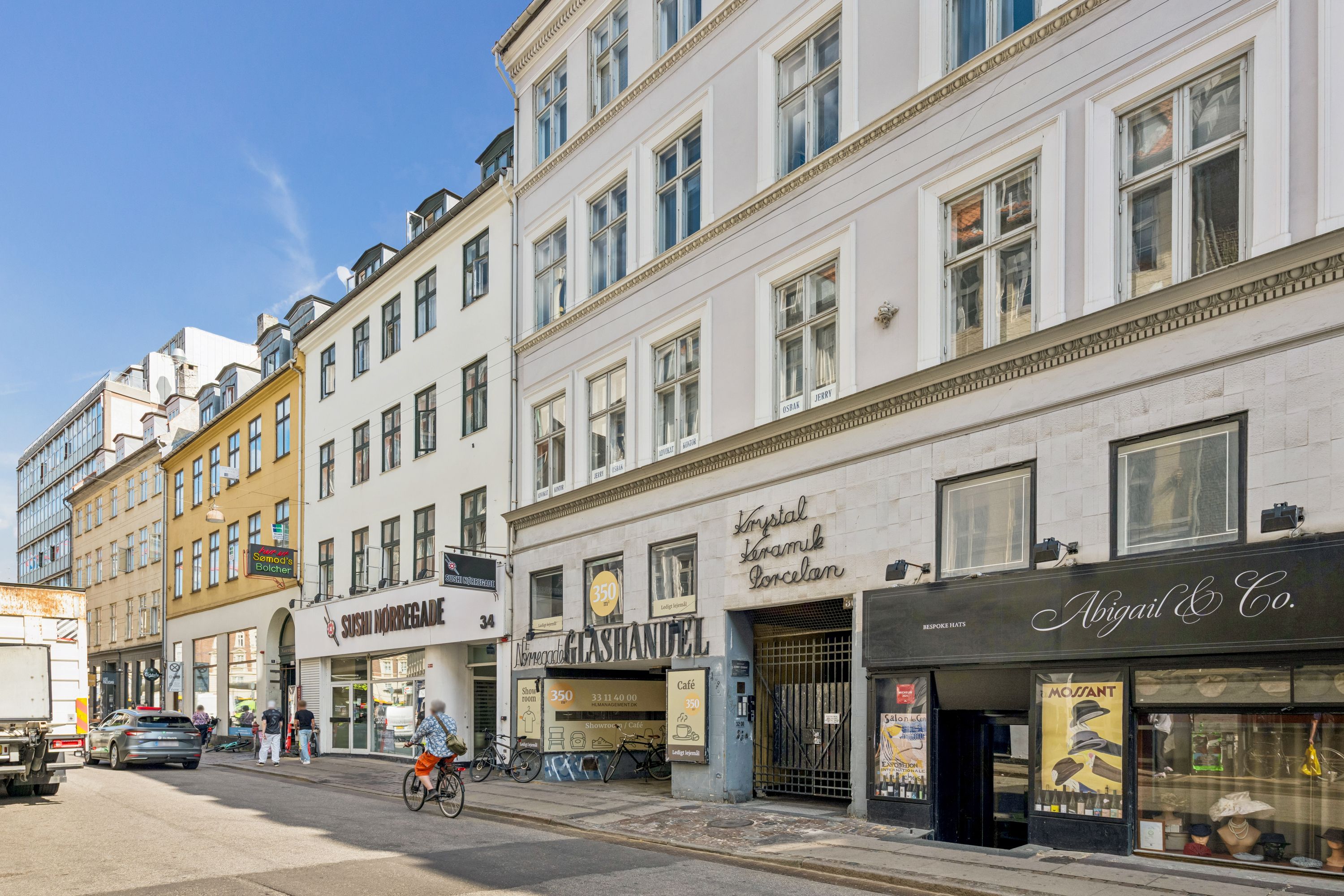En cykelkører passerer en stor bygning på Nørregade 30, hvor der er butikker og caféer som Abigail & Co og Café Nimegced. En bil kører langs vejen og et nyt vogn er parkeret på siden