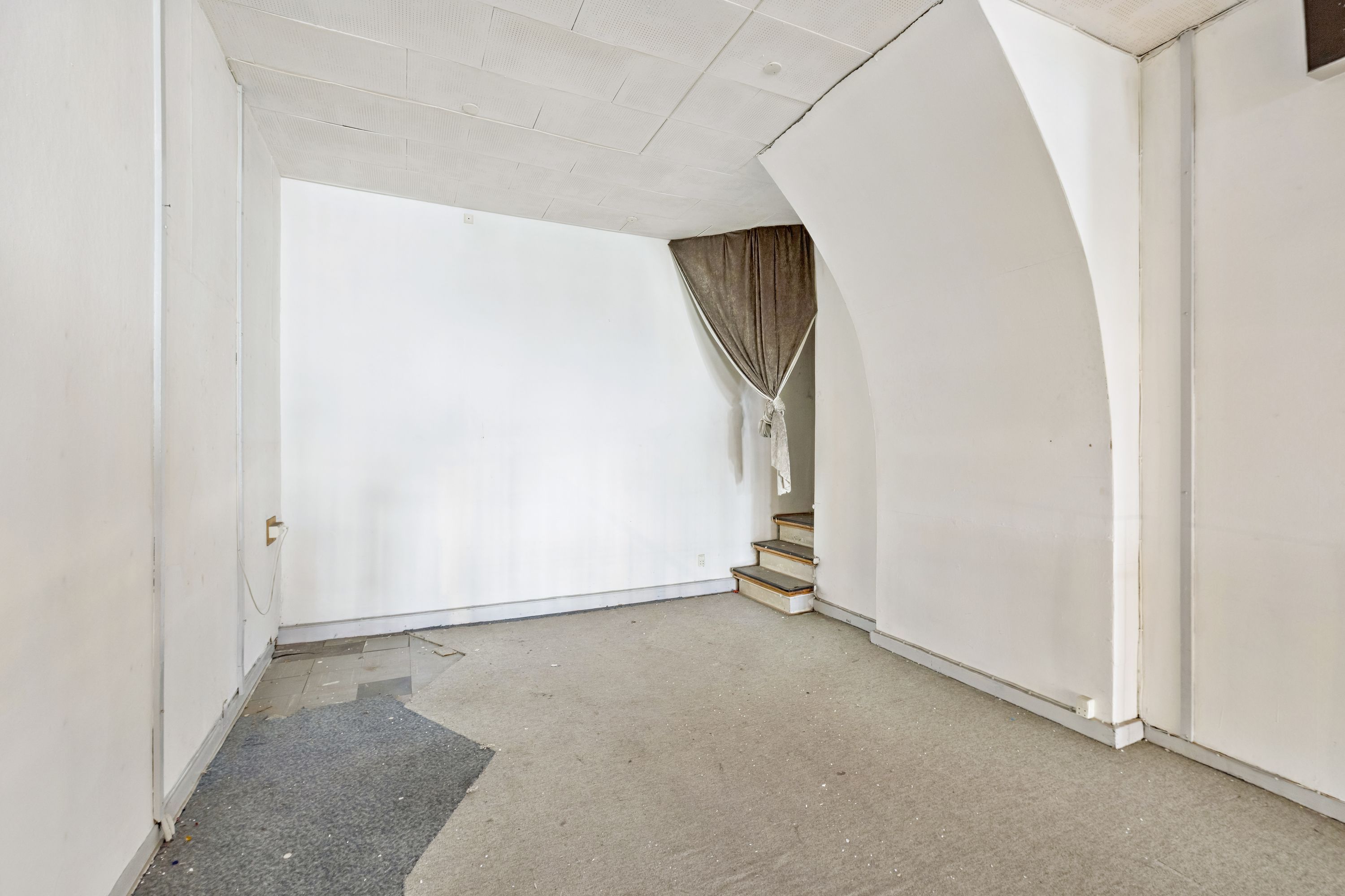 En stor og lys stue med hvidmalede vægge og en høj loftshøjde samt et stykke gulvtæppe og trætrapper til kælderen